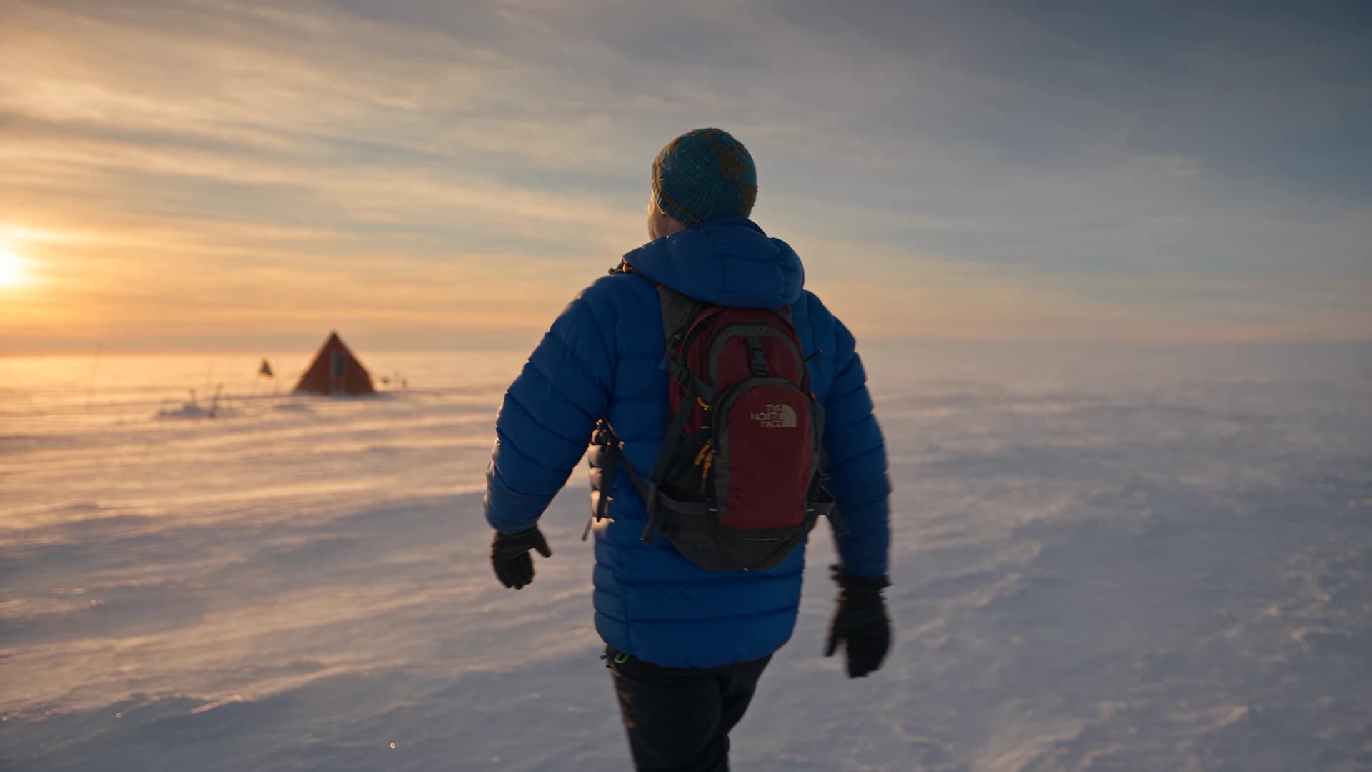 A glaciologist in a blue down jacket crosses a polar ice sheet toward a tent at sunset on the Grounding Line expedition